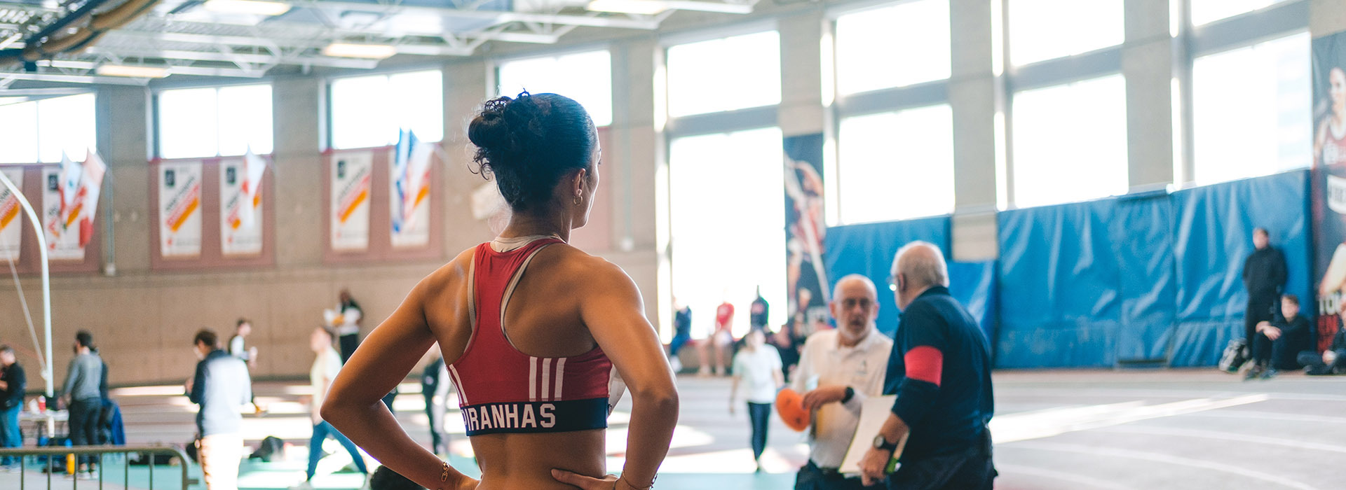 Une athlète observe attentivement une compétition de lutte, entourée de spectateurs et d'entraîneurs dans un espace dynamique.