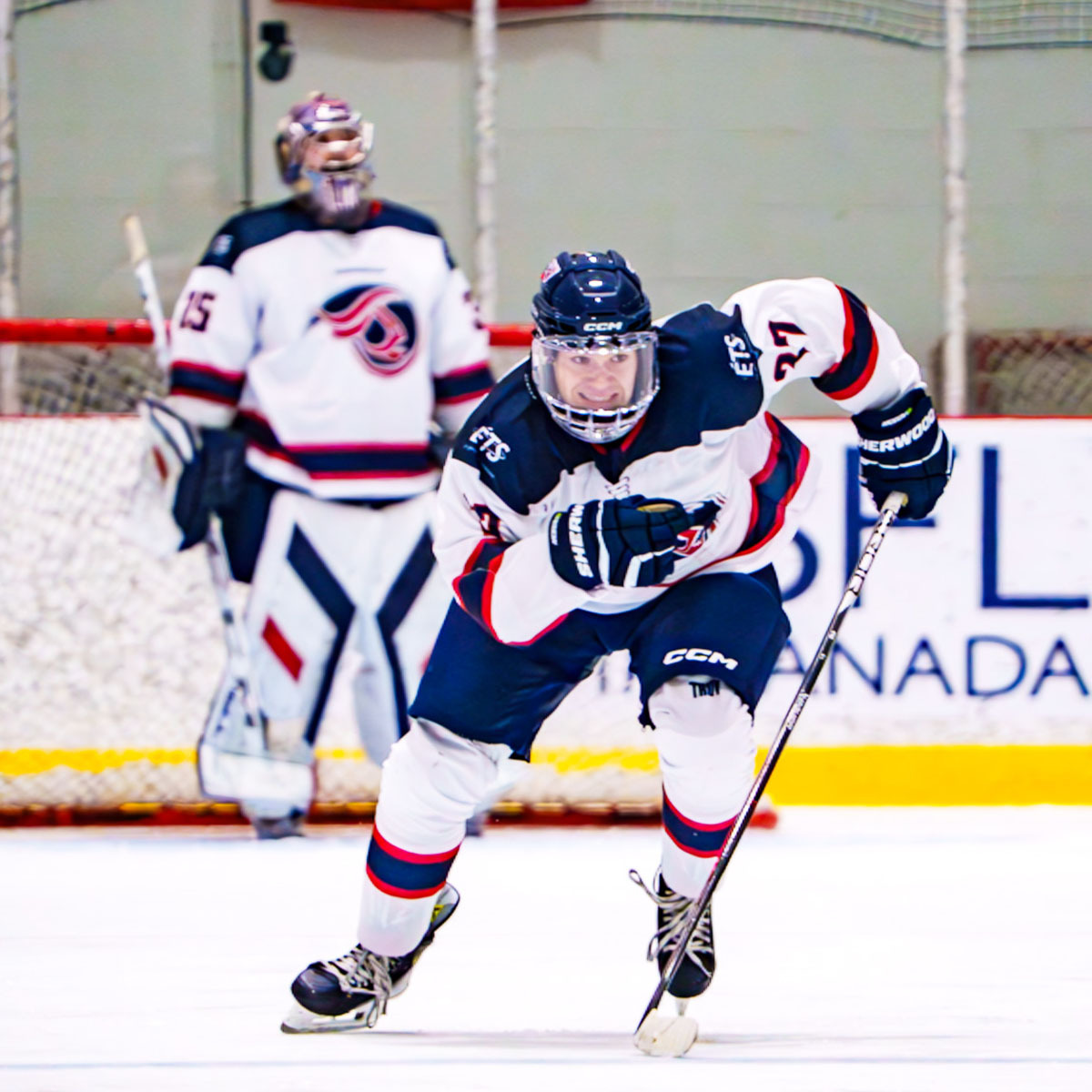 Un joueur de hockey scrute le jeu sur la glace, prêt à foncer vers le but, tandis que son gardien de but veille en arrière-plan.