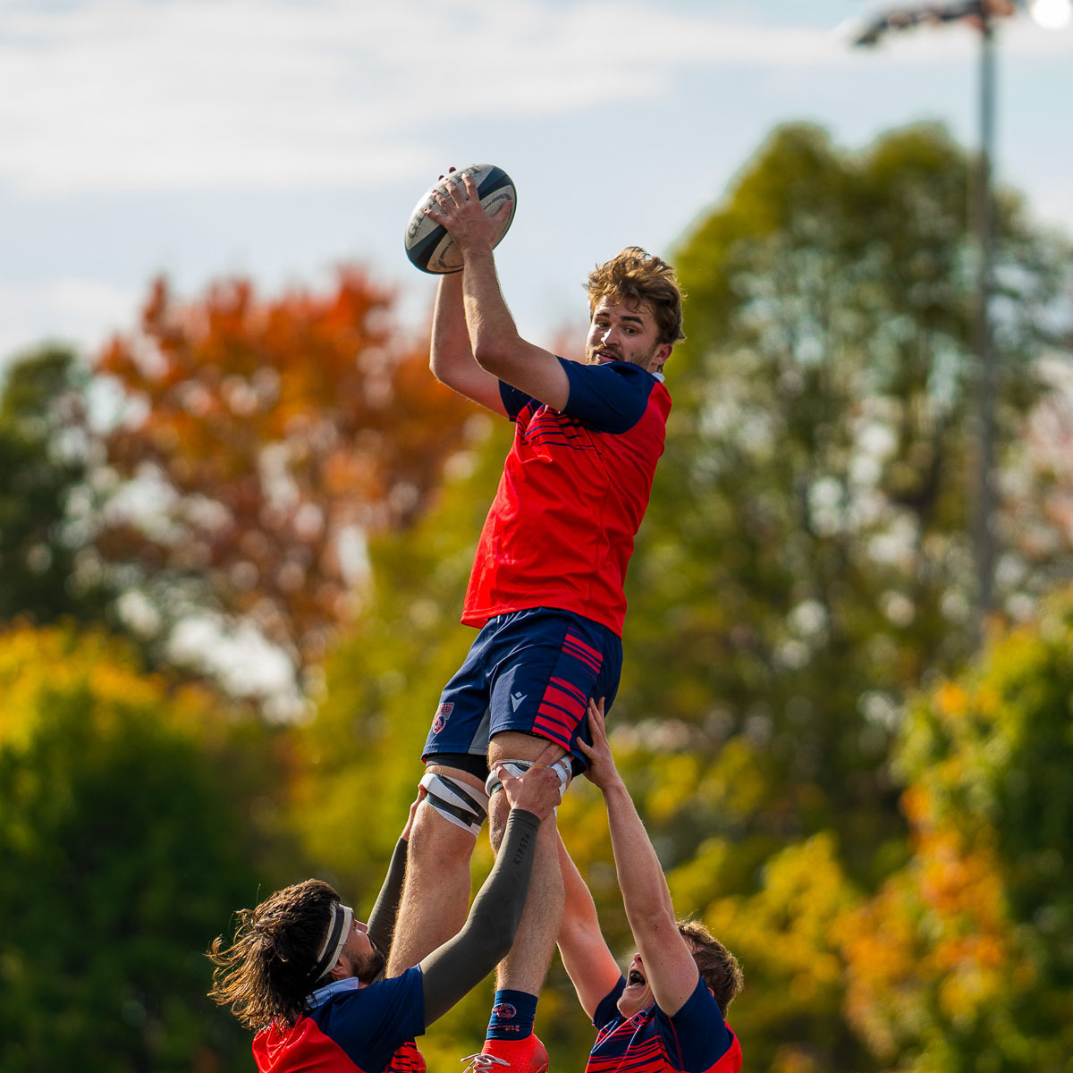 Un joueur de rugby en l'air, soutenu par deux coéquipiers, saisit le ballon sous un ciel dégagé et des arbres aux couleurs d'automne.