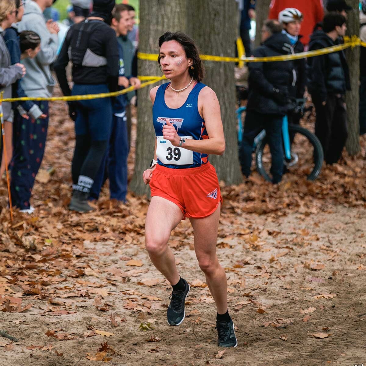 Une athlète s'élance sur un parcours de course à pied, entourée de supporters dans un cadre automnal. L'énergie et l'engagement sont palpables.