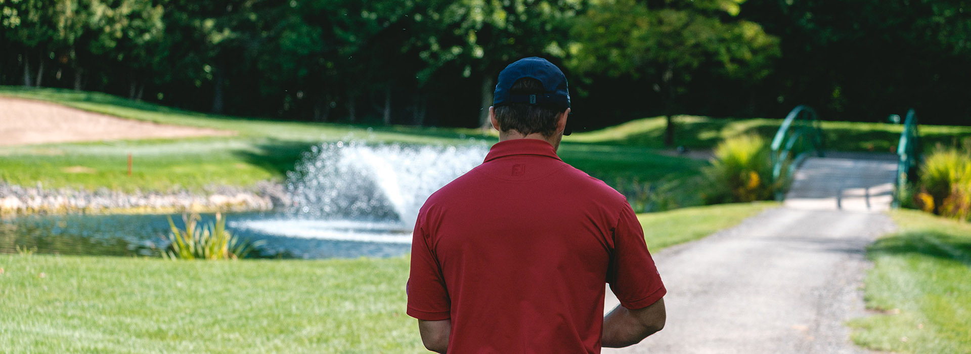 Un homme en polo rouge marche le long d'un chemin, avec un environ verdoyant et un étang en arrière-plan, au calme d'un parcours de golf.