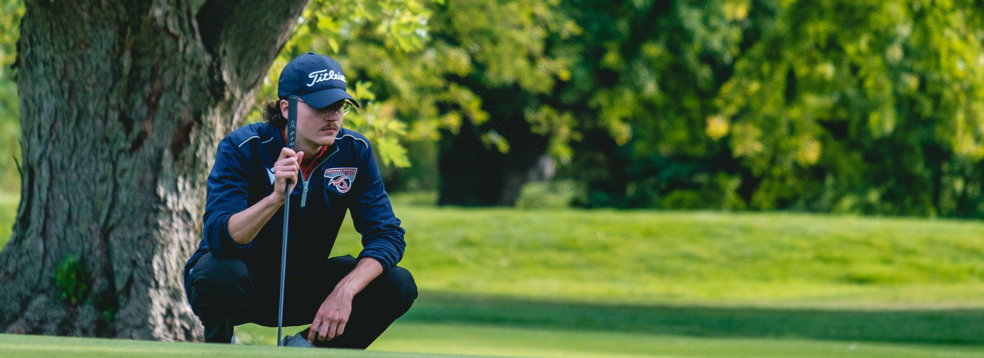 A student focuses intently while preparing to putt on a lush green golf course, surrounded by trees in a serene setting.