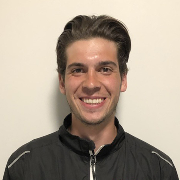 A cheerful young man with wavy hair, wearing a black zip-up jacket, smiles against a light background.