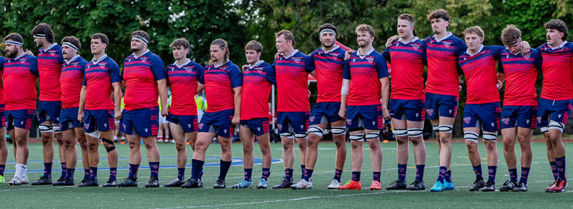 A rugby team stands in formation, wearing vibrant red and blue uniforms, ready for an upcoming match on a green field.