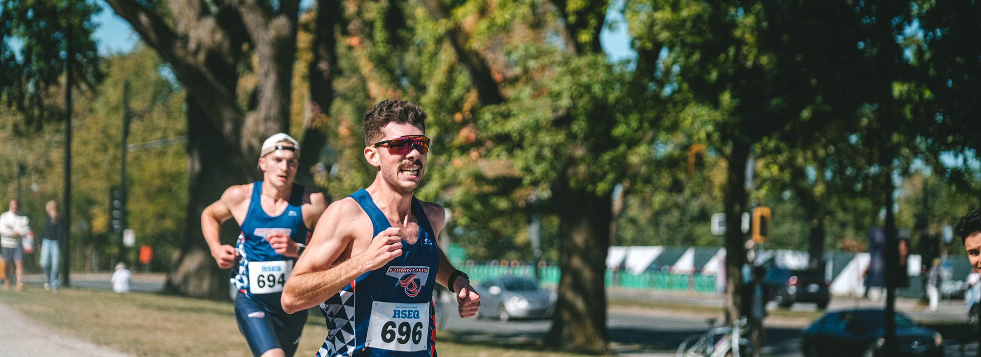 Two male runners compete during a race, showcasing determination and athleticism against a backdrop of trees and spectators.