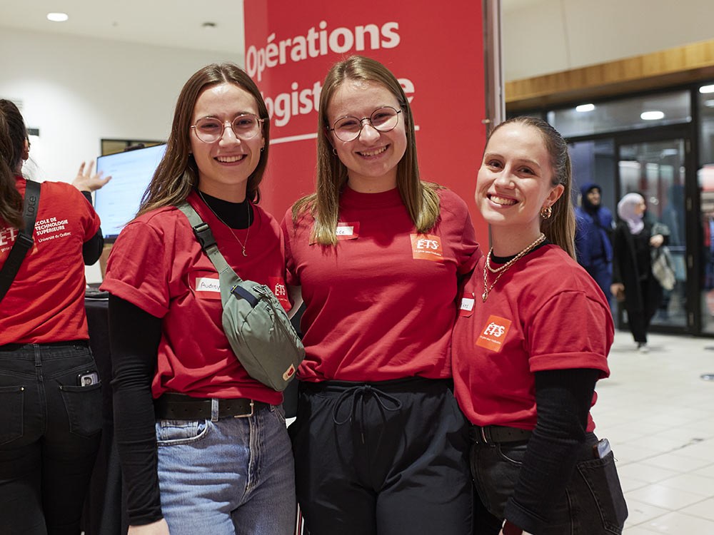 Trois étudiantes souriantes portent des t-shirts rouges, représentant l'ÉTS lors d'un événement d'accueil. Ambiance conviviale et dynamique.