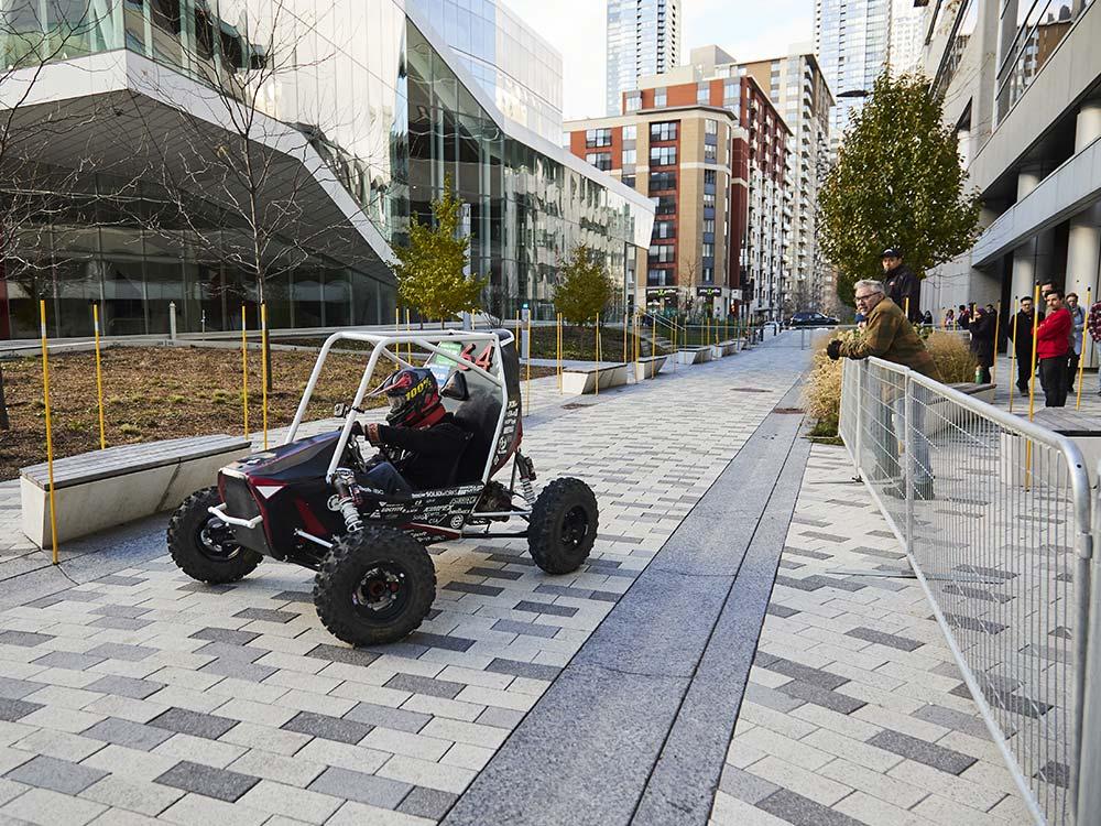 Une buggy innovant roule sur un chemin urbain, attirant l'attention des passants, au cœur d'un campus moderne.