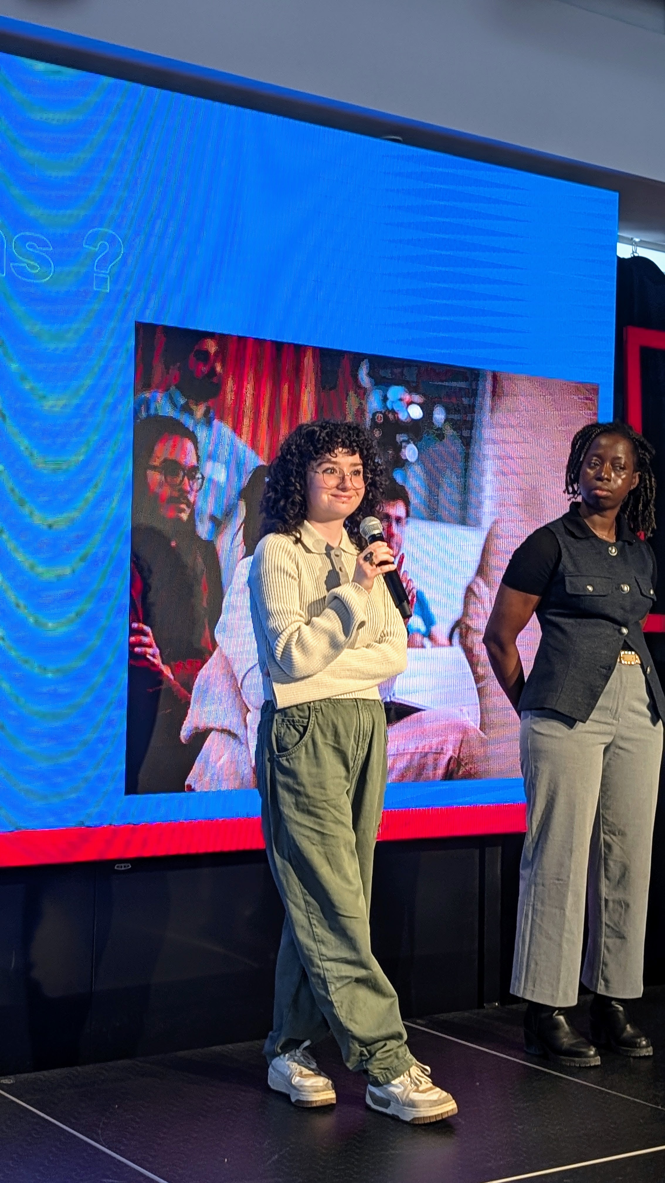 Deux intervenantes s'expriment sur une scène face à un auditoire, avec un grand écran en arrière-plan. Ambiance dynamique et engageante.