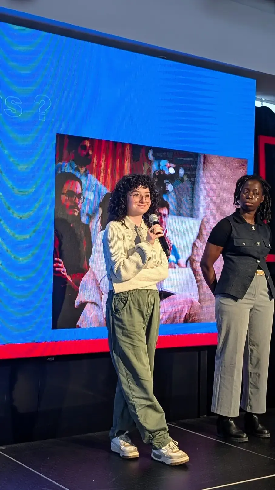 Deux intervenantes s'expriment sur une scène face à un auditoire, avec un grand écran en arrière-plan. Ambiance dynamique et engageante.