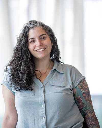 Femme souriante au look décontracté, cheveux longs et bouclés, vêtue d'une chemise claire, avec des tatouages artistiques sur le bras.