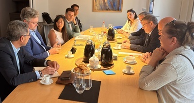 Réunion autour d'une grande table avec des participants discutant, des tasses de café et des croissants disposés. Ambiance collaborative et professionnelle.