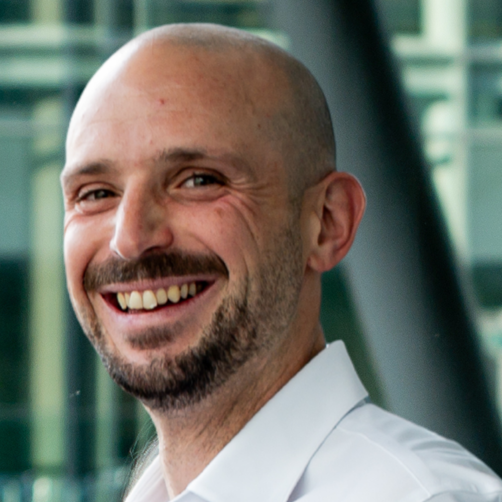A smiling individual in a white shirt, set against a modern technological backdrop, exudes a positive and approachable presence.