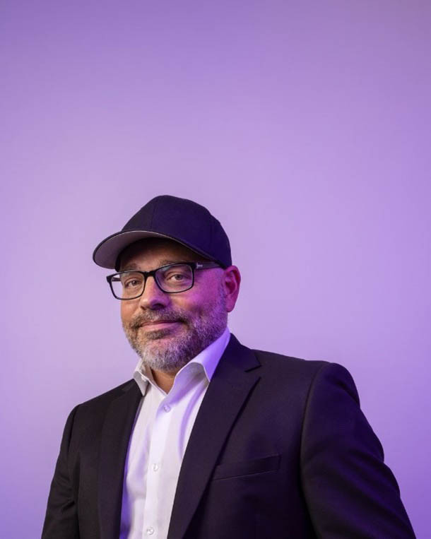 Personne souriante portant une casquette et des lunettes, habillée en costume noir, devant un fond violet. Ambiance professionnelle et moderne.