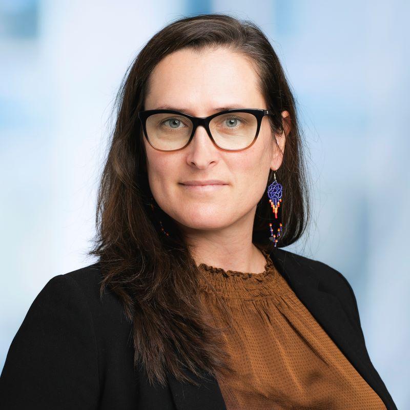 Femme souriante, lunettes épaisses, portant des boucles d'oreilles colorées et une blouse en tissu texturé sous un blazer noir.