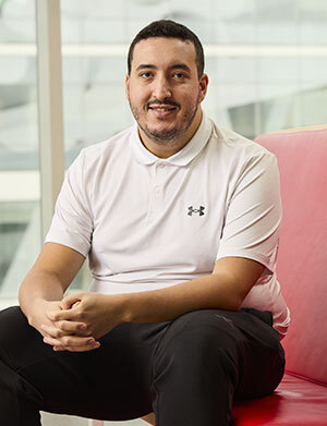 Un étudiant souriant porte un polo blanc, assis sur un canapé rouge, dans un environnement moderne et lumineux.