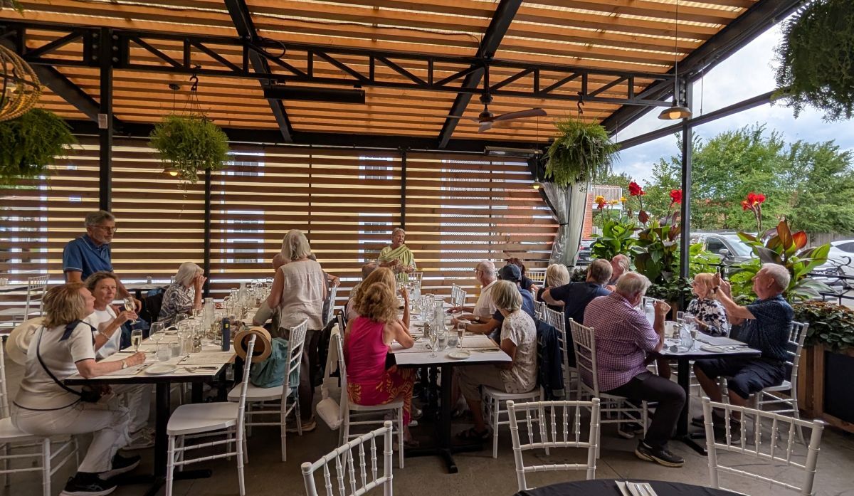 Un groupe de personnes se retrouve autour de tables dans une ambiance conviviale, profitant d'un repas entouré de verdure.