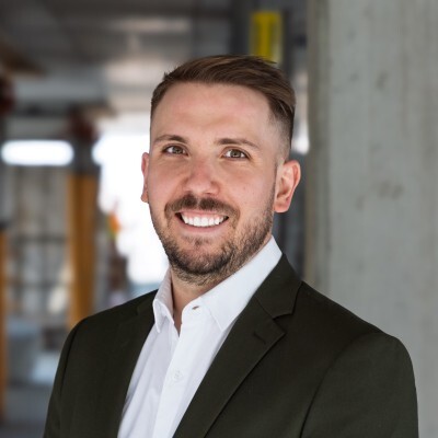 Un professionnel souriant, vêtu d'un blazer et d'une chemise blanche, se tient dans un environnement moderne et éclairé.