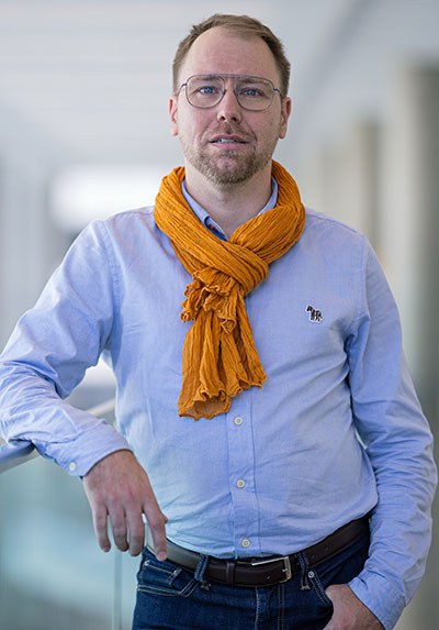 Un homme souriant porte une chemise bleue et une écharpe orange, se tenant près d'une balustrade dans un environnement moderne.