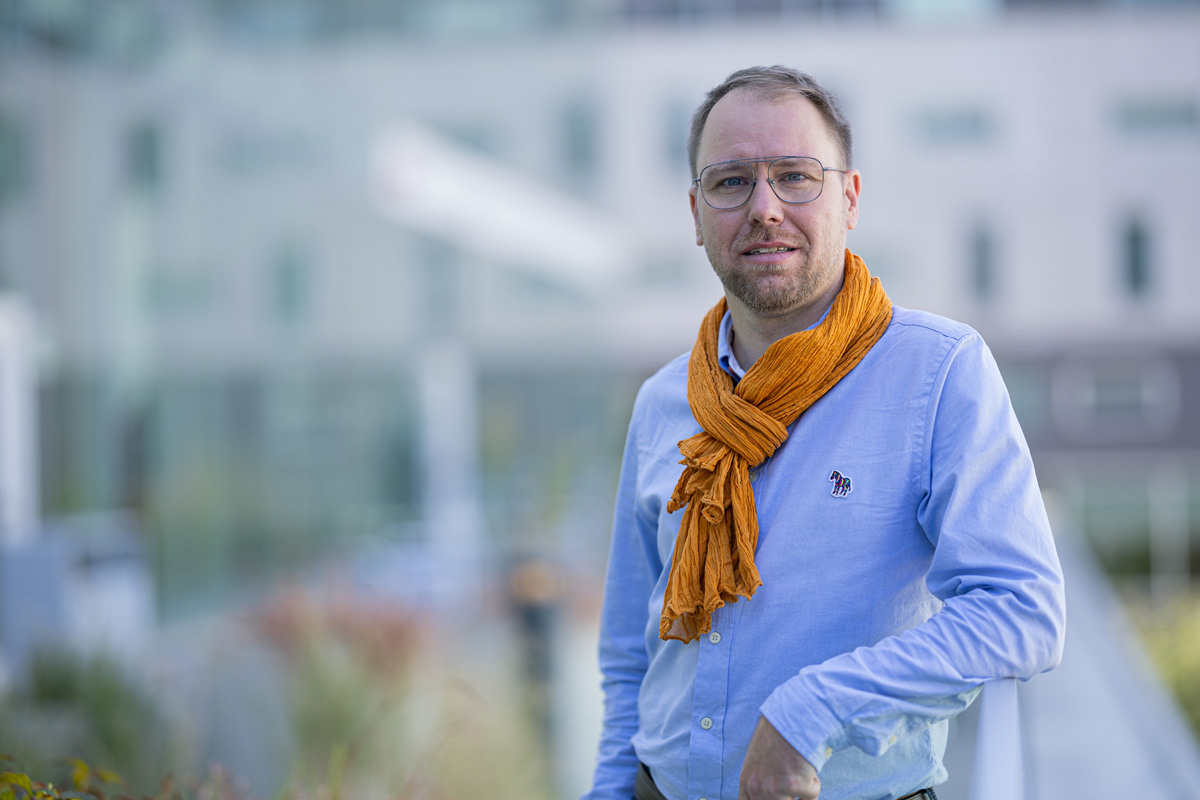 Un professionnel souriant porte une chemise bleue et une écharpe orange, se tenant dans un cadre moderne et lumineux.