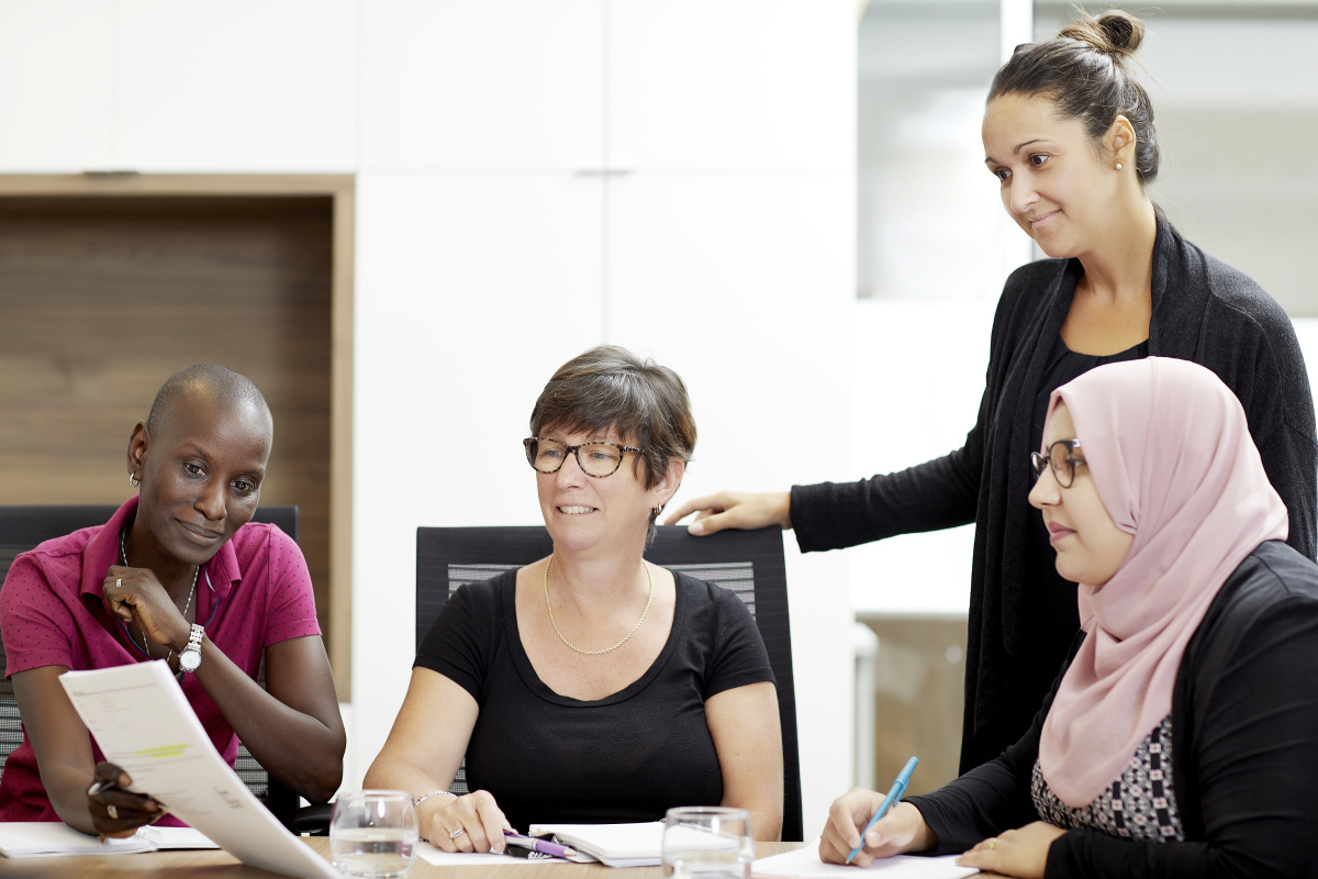 Quatre femmes en réunion échangent des idées, chacune avec un document, apportant des contributions variées dans un cadre collaboratif.