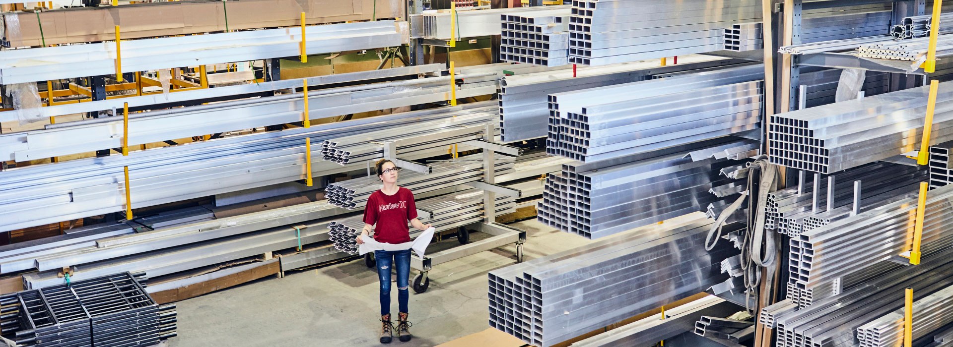 Étudiante en ingénierie dans un environnement industriel, entourée de matériaux en aluminium, examinant des plans de projet.