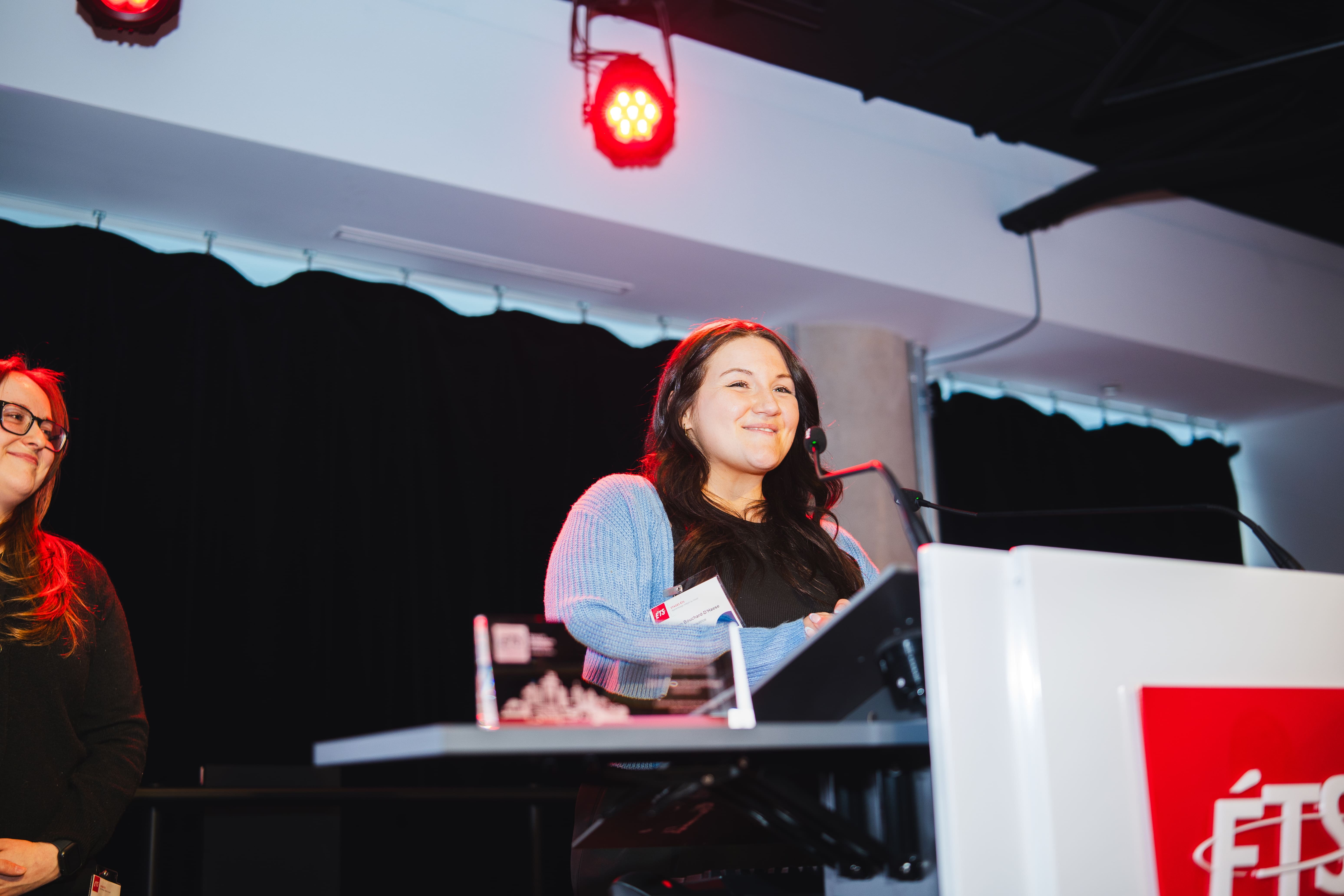 Une intervenante souriante s’apprête à prendre la parole lors d’un événement, tandis qu'une collègue l’écoute attentivement.