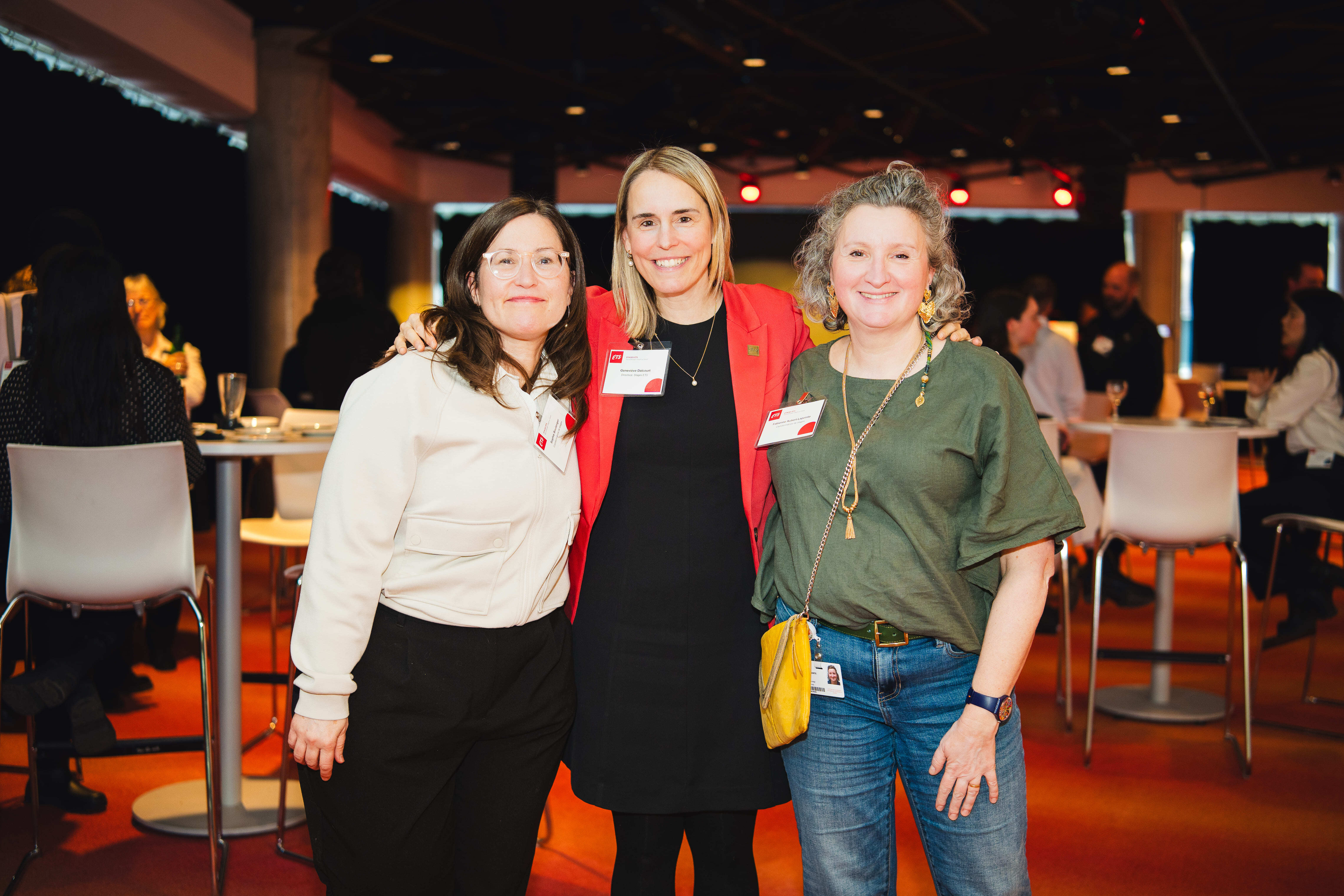 Trois femmes souriantes posent ensemble lors d'un événement, avec un fond animé et des tables, créant une ambiance conviviale et professionnelle.