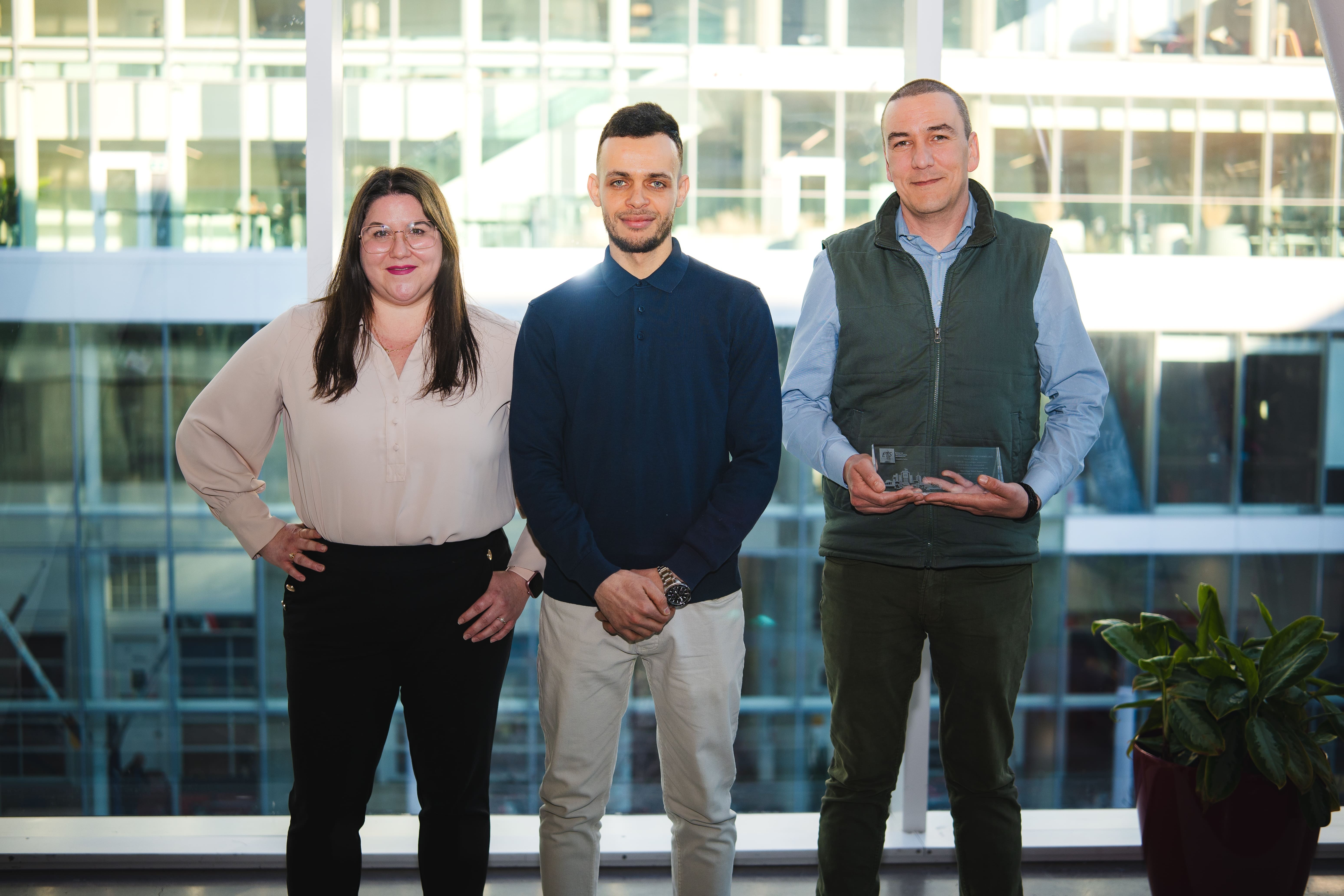 Trois personnes se tiennent côte à côte, souriantes, dans un cadre lumineux avec des fenêtres modernes en arrière-plan.