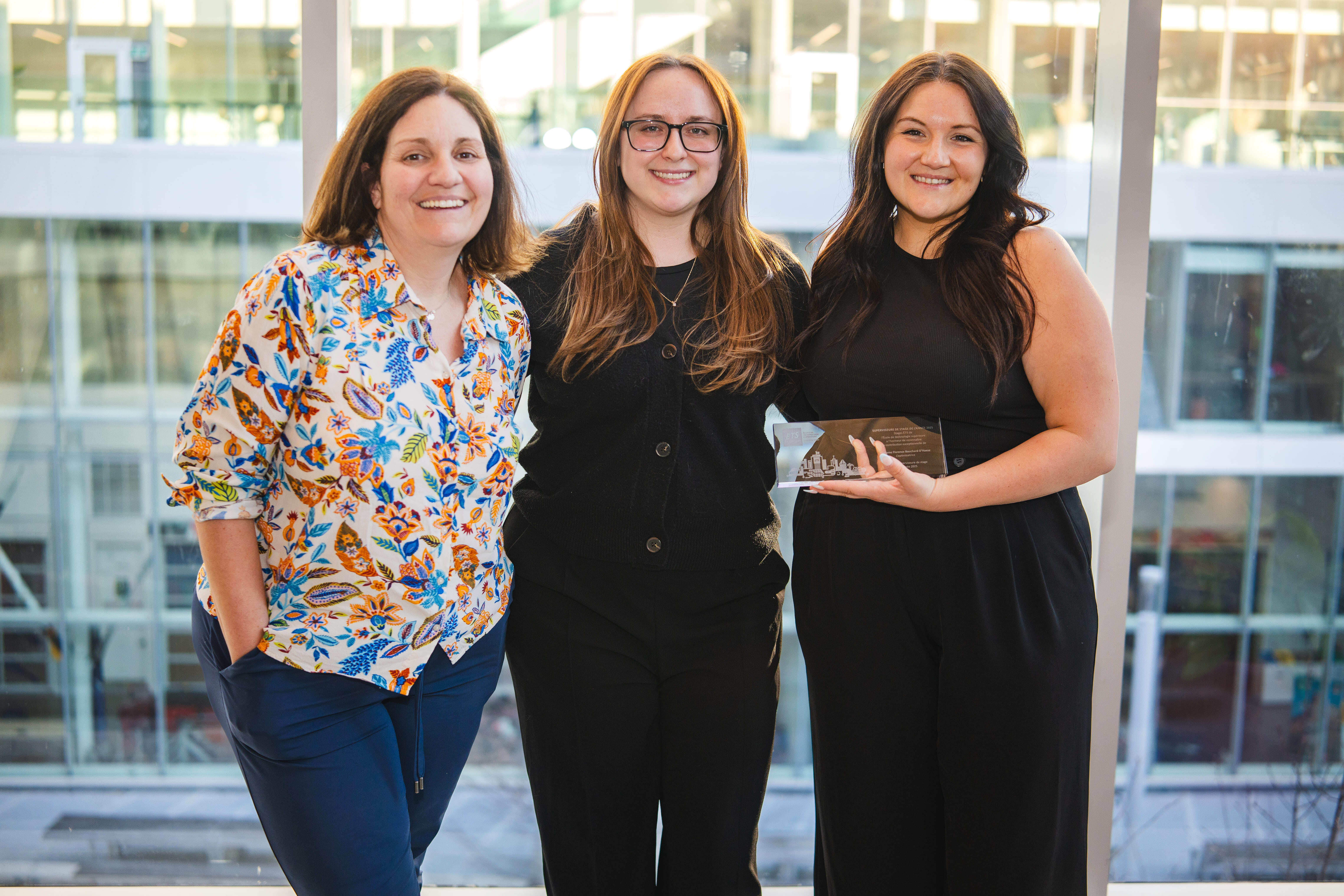 Trois femmes souriantes posent ensemble, tenant un prix, avec un fond lumineux et moderne d’un environnement universitaire.