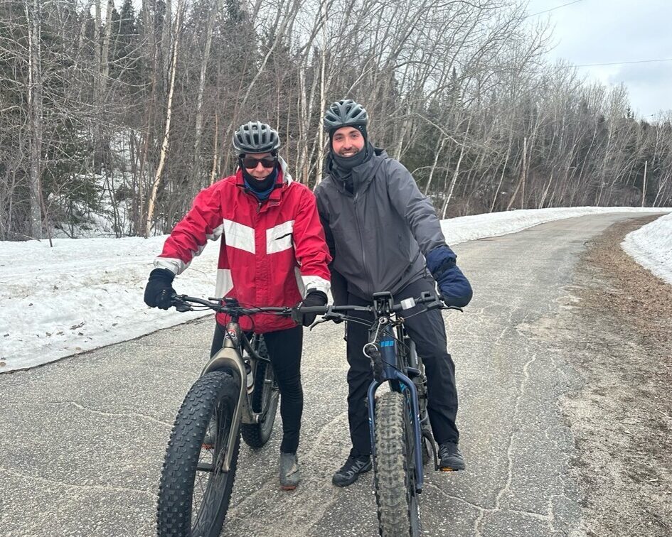 Deux cyclistes souriants prennent une pause sur une route enneigée, profitant d'une journée d'hiver en plein air.