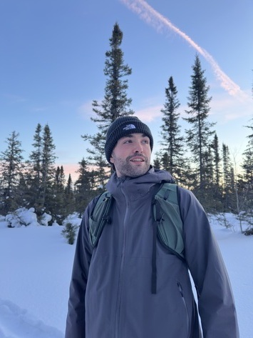 Un homme en tenue d'hiver se tient dans un paysage enneigé, entouré de sapins, sous un ciel clair au lever du jour.