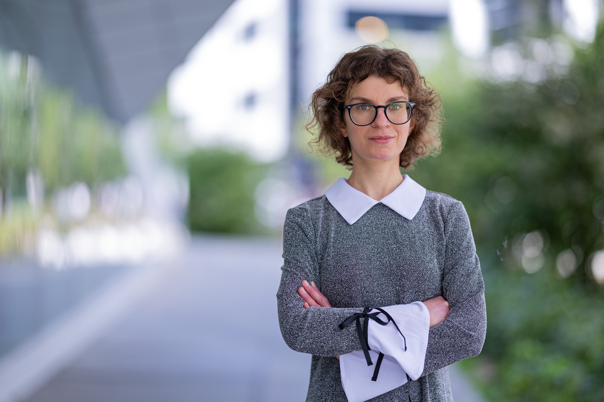 A smiling woman, elegantly dressed, wearing glasses, stands in a modern and green environment.