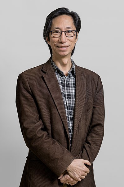 Un professeur souriant, vêtu d'un blazer marron et d'une chemise à carreaux, se tient avec confiance devant un fond clair.