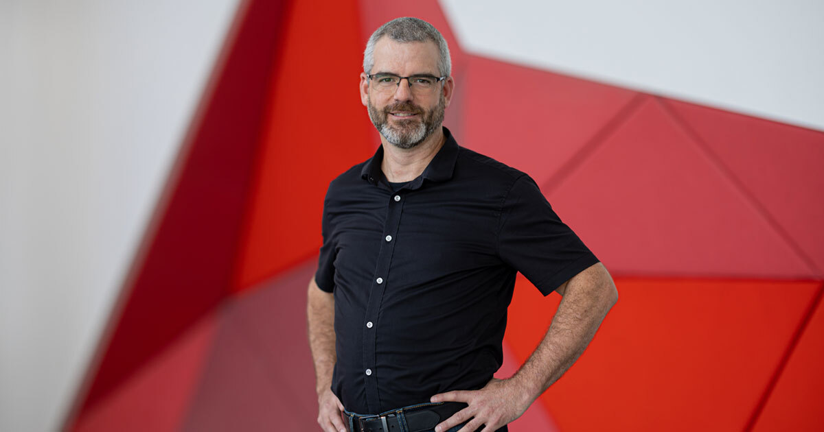 A professional standing confidently in front of a modern, red and white geometric background, showcasing a contemporary academic setting.