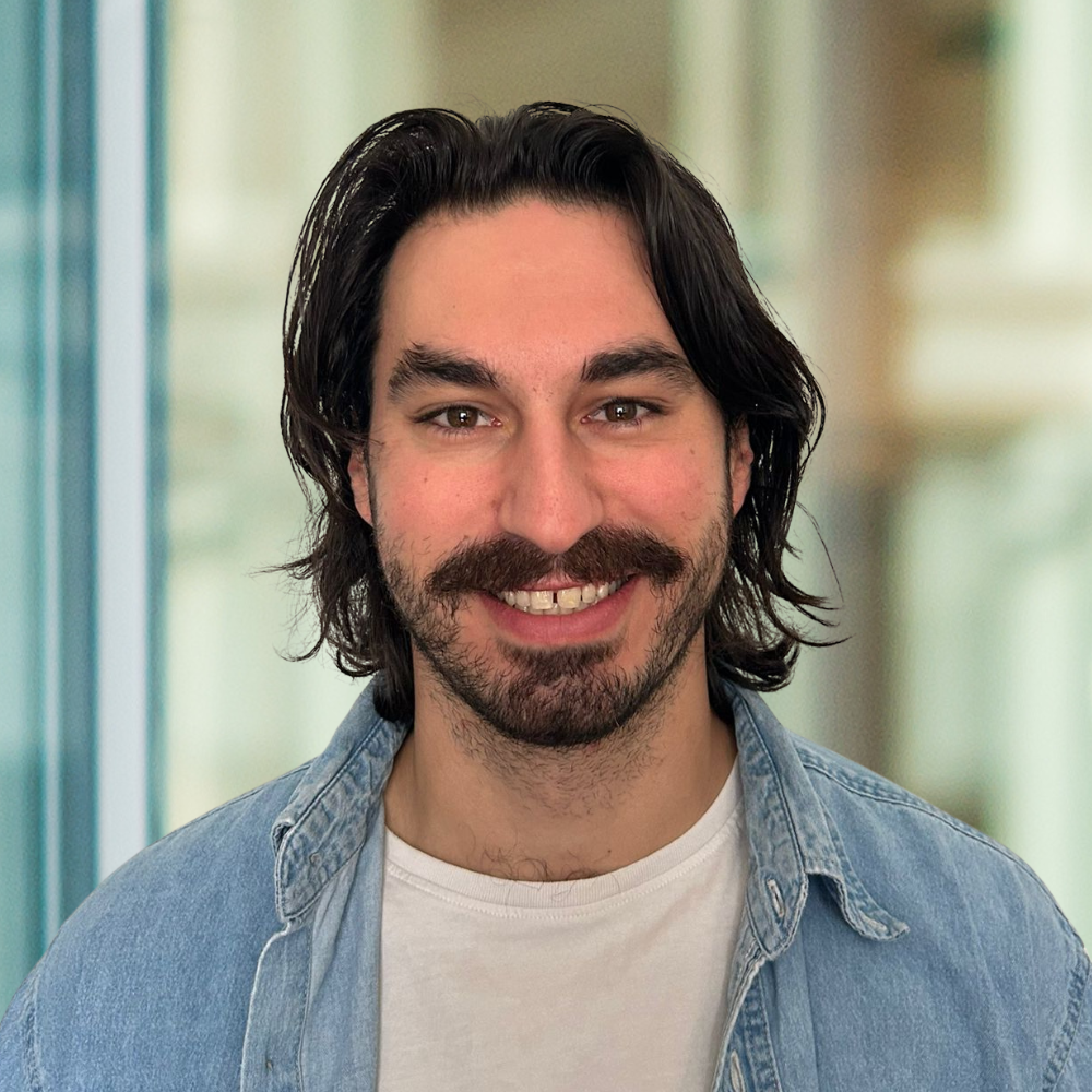 A smiling student with dark, long hair and a mustache, wearing a denim jacket over a white t-shirt, in a bright, modern setting.