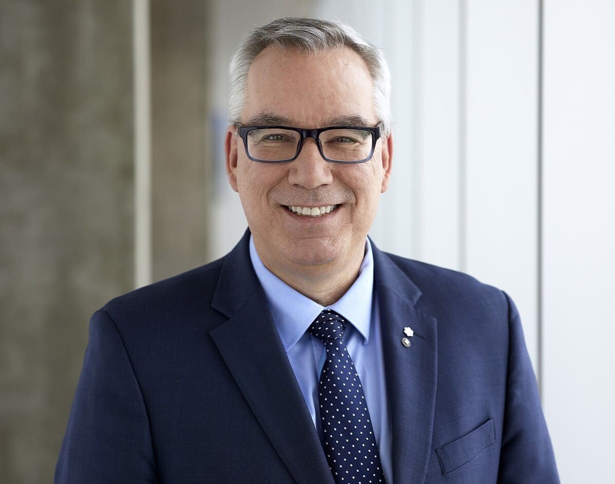Un homme souriant, en costume, avec des lunettes, se tient dans un environnement moderne et lumineux, reflétant professionnalisme et accessibilité.