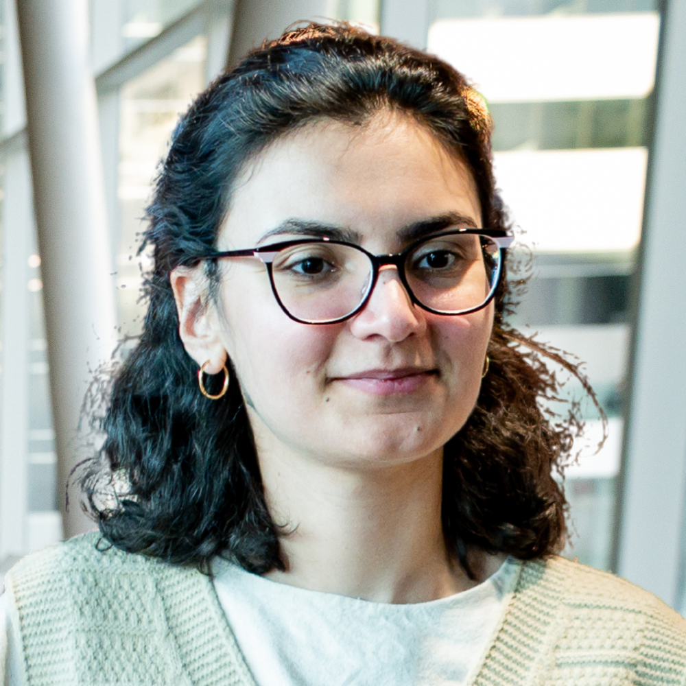A student with curly hair and glasses smiles confidently, showcasing a vibrant academic environment in a modern university setting.